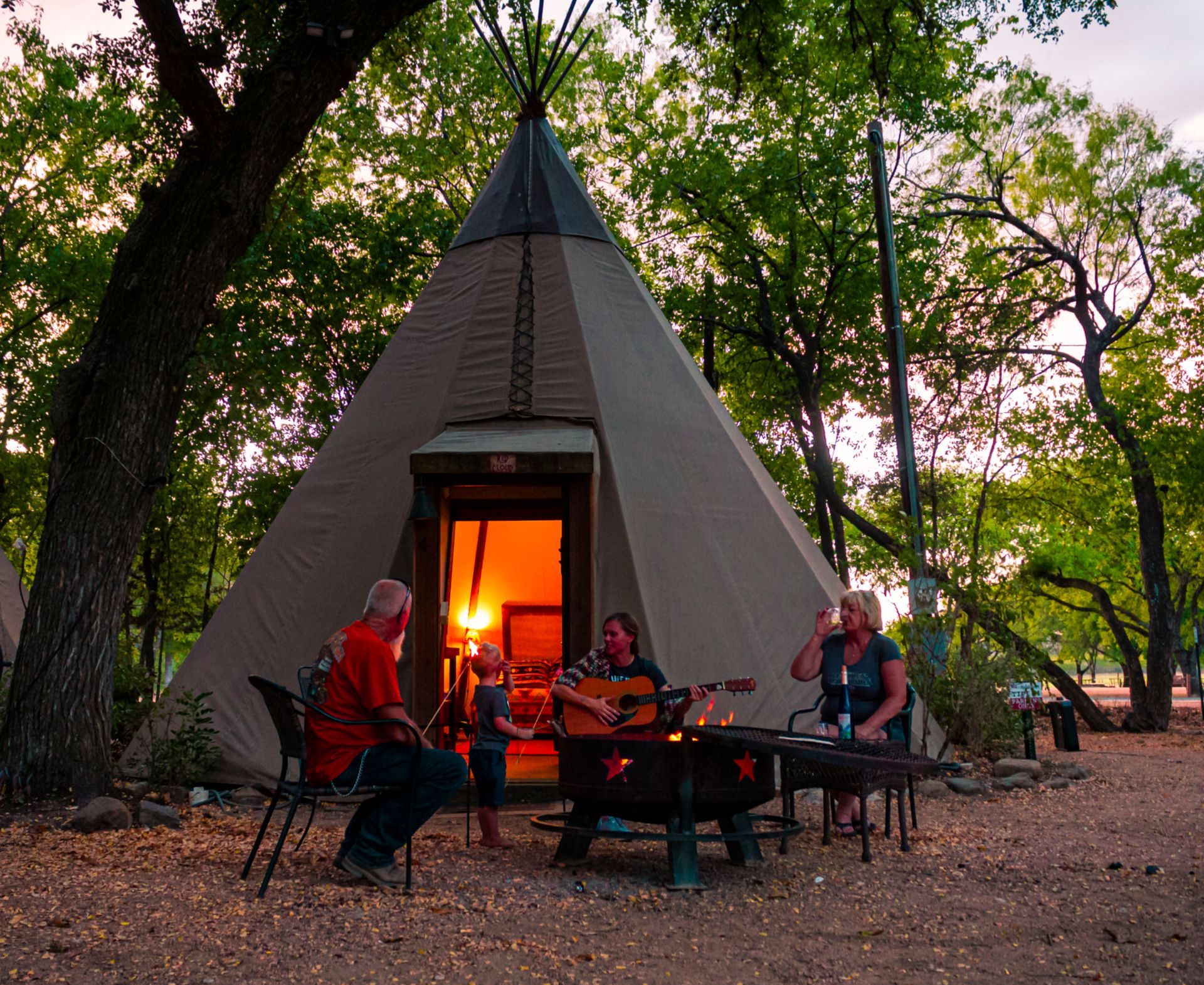 People around a campfire in front of a Tipi, guitar player, evening, trees.