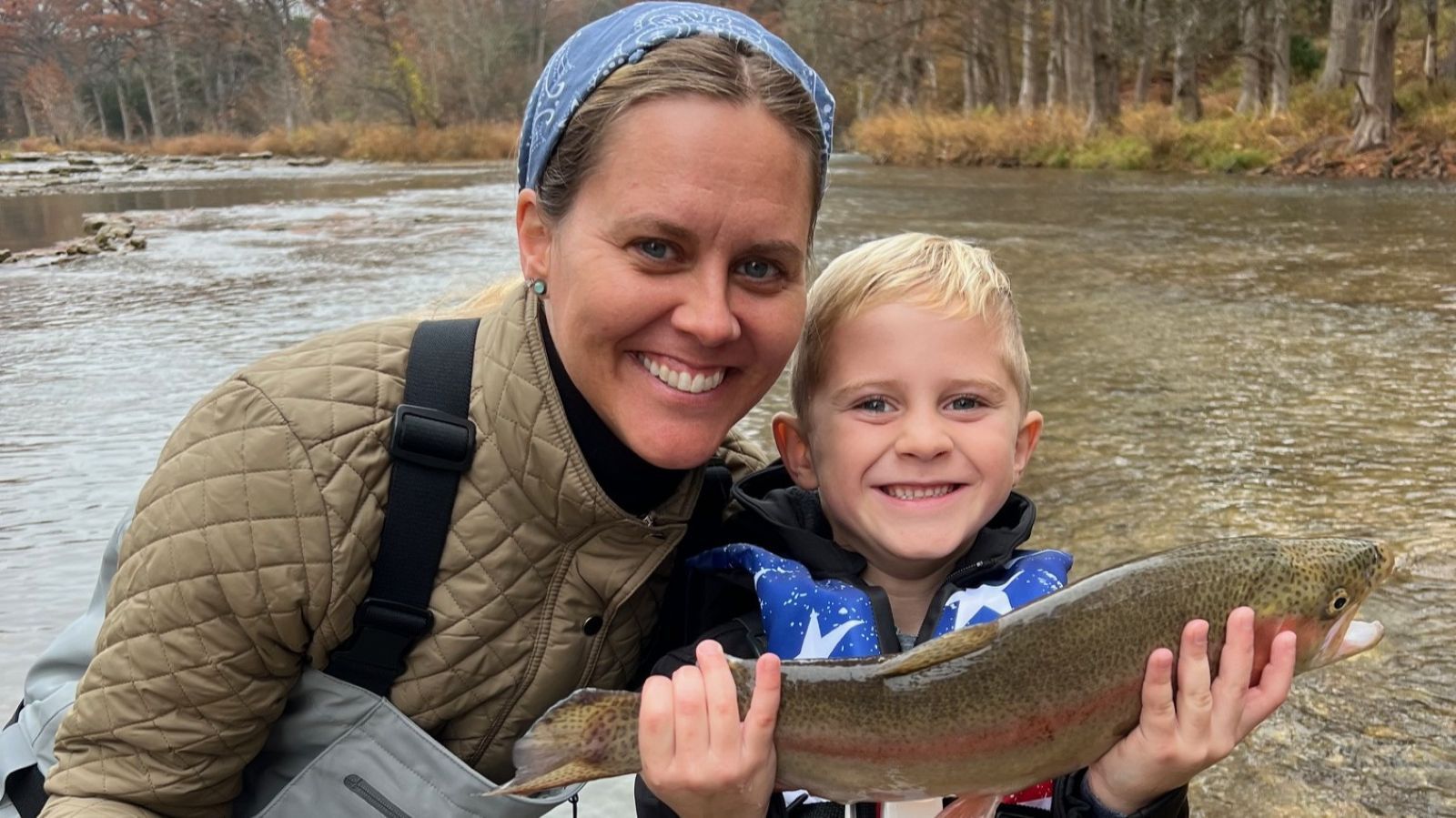 Woman and boy smiling, holding a large fish near a river.