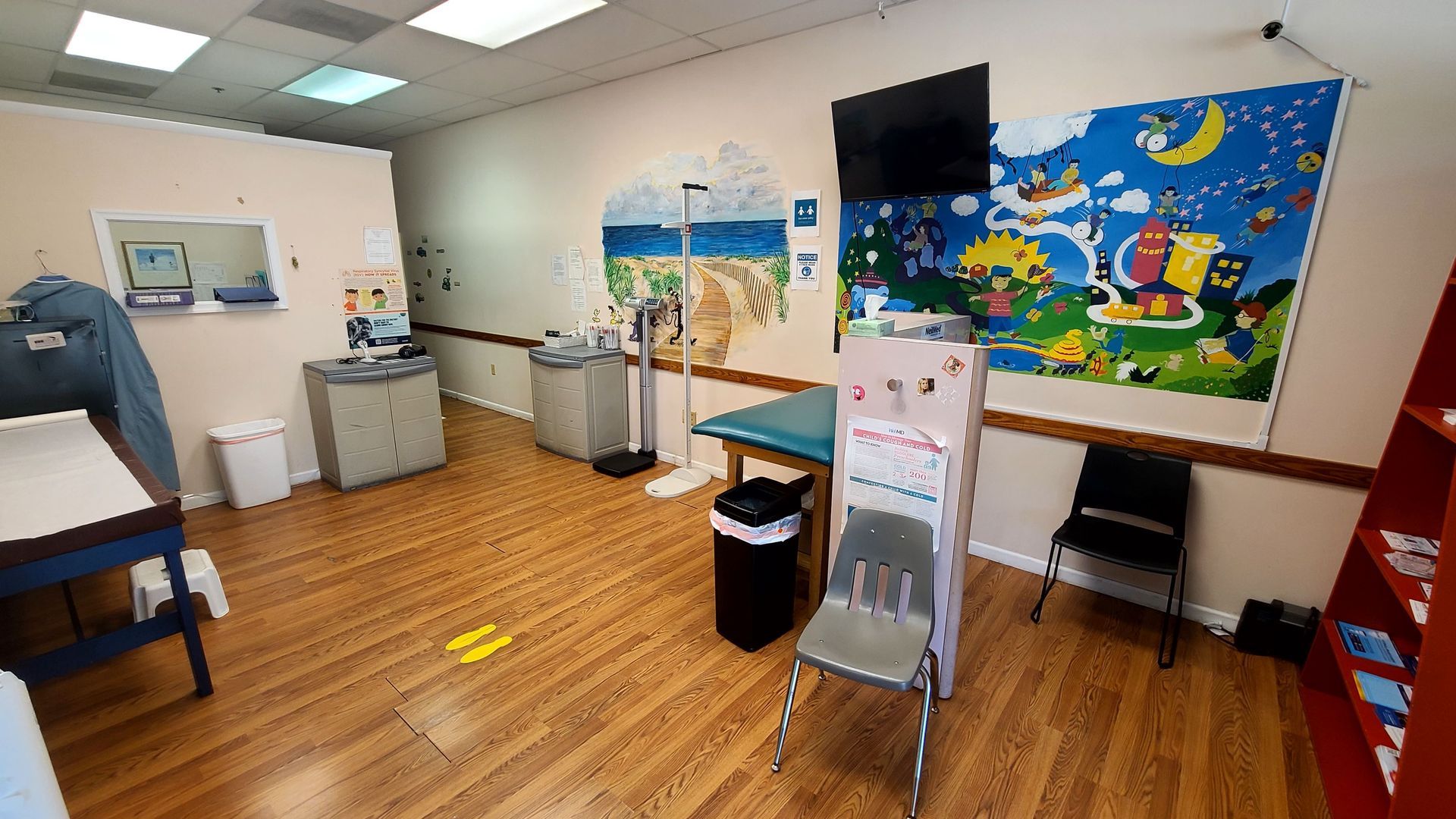Pediatrician's office exam room with colorful mural, medical equipment, examination table, and two chairs.