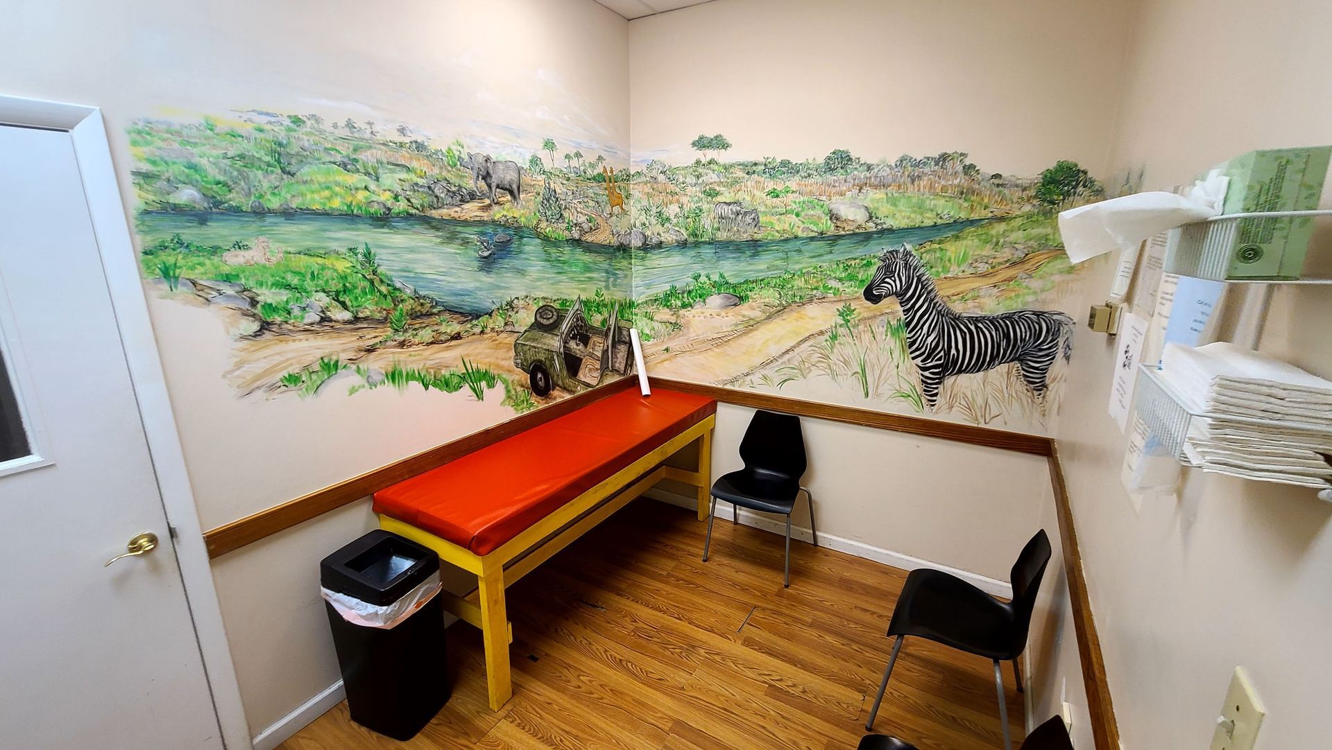 Examination room with a mural of a savanna scene, red examination table, and black chairs.