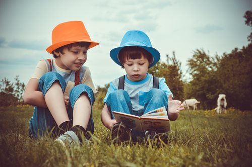 Two children in hats reading a book together in a grassy field.