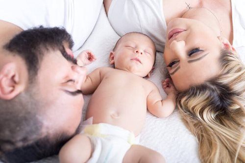 Parents and newborn baby lying together on a white bed, all asleep.
