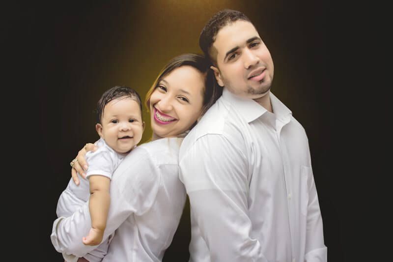 Family of three in white shirts, smiling against a dark background. Baby held by mother.