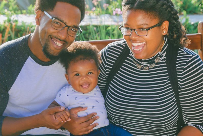 Family of three smiles outdoors; parents hold a baby.