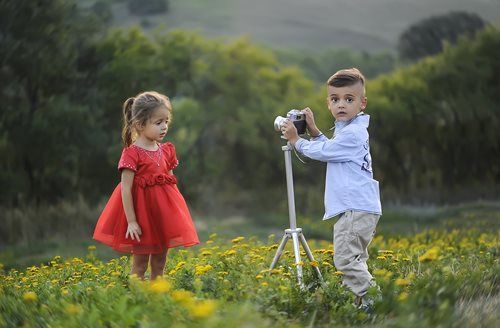 Boy using camera on tripod, photographing girl in red dress in a field of yellow flowers.