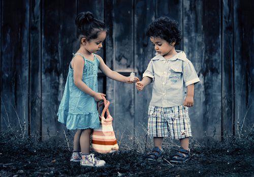 Girl in blue dress giving something to boy in shorts, standing in front of a wooden wall.