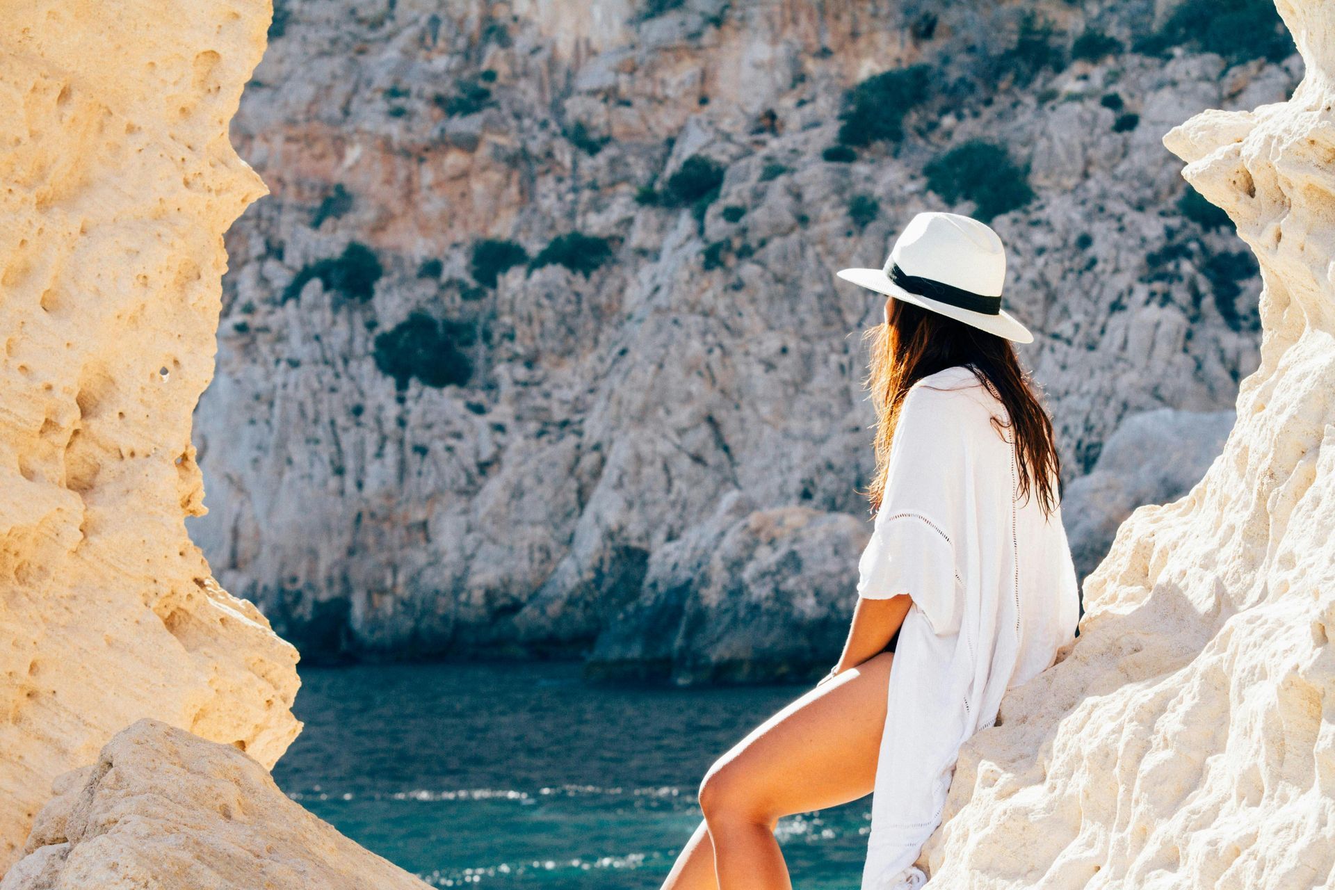 A woman in a white hat is sitting on a rock near the ocean.