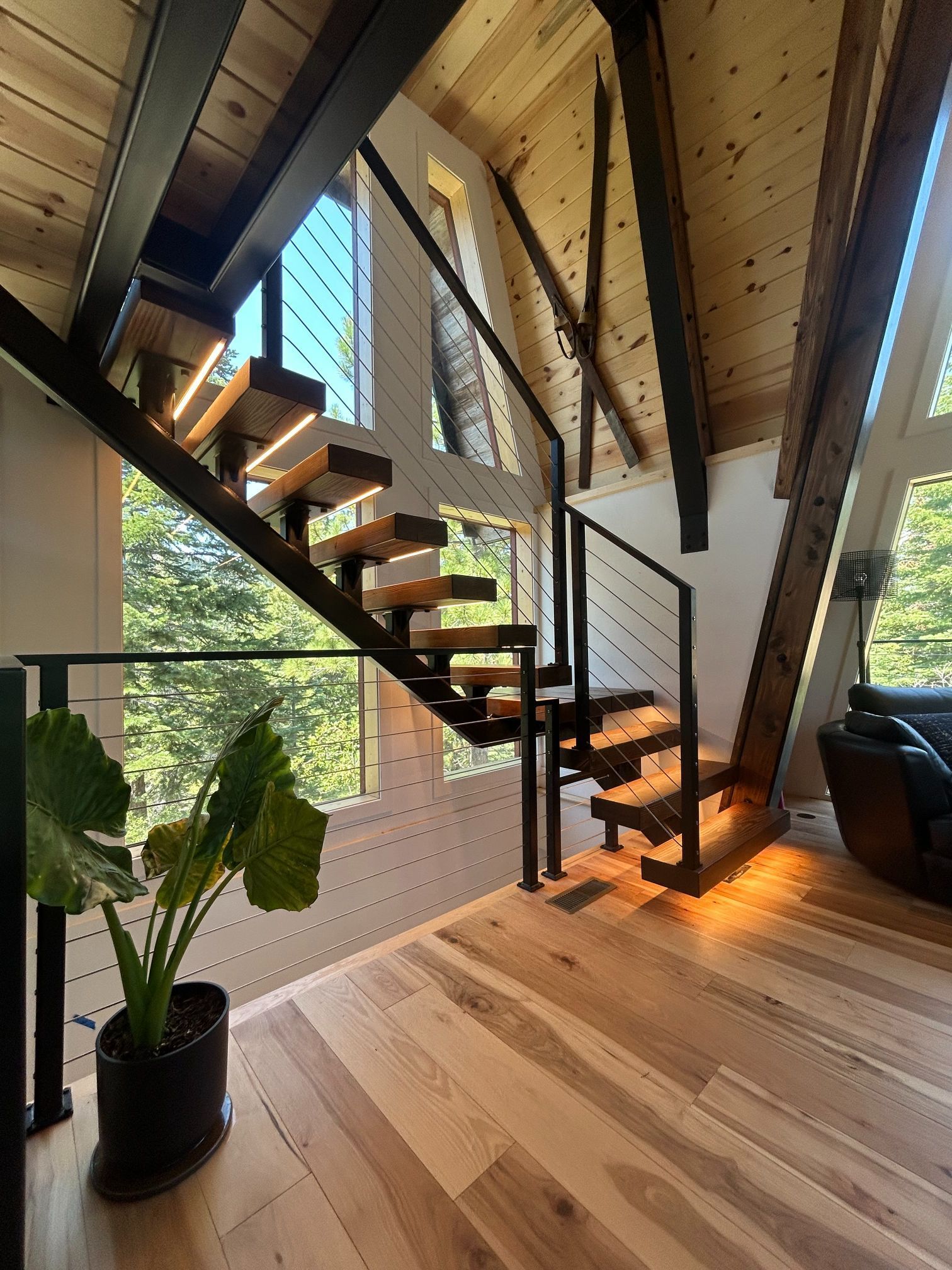 Wooden staircase with black metal railing, leading to a second story, with large windows overlooking trees.