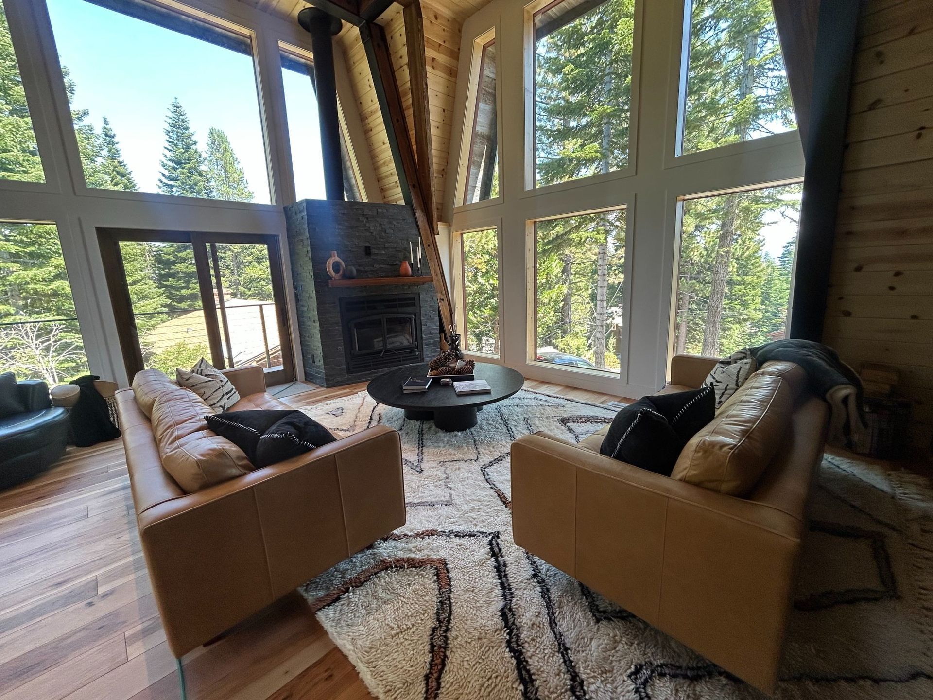 Living room with large windows overlooking a forest. Two brown leather couches face a fireplace.