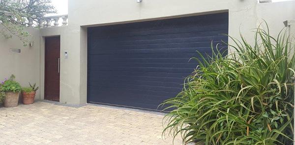 Dark blue garage door with adjacent door, plants and paved driveway.