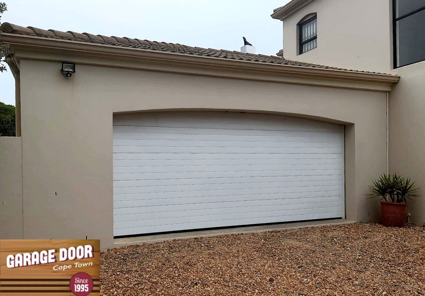 Tan garage with white roll-up door, gravel driveway. Brown roof and potted plant.