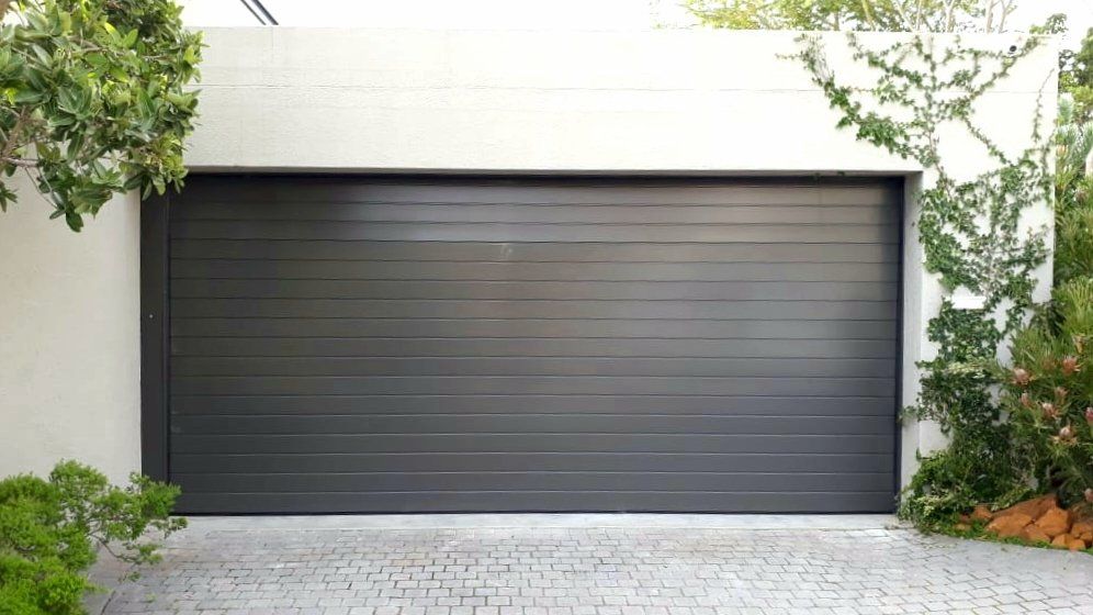 Gray garage door on a white building, surrounded by greenery and a stone driveway.