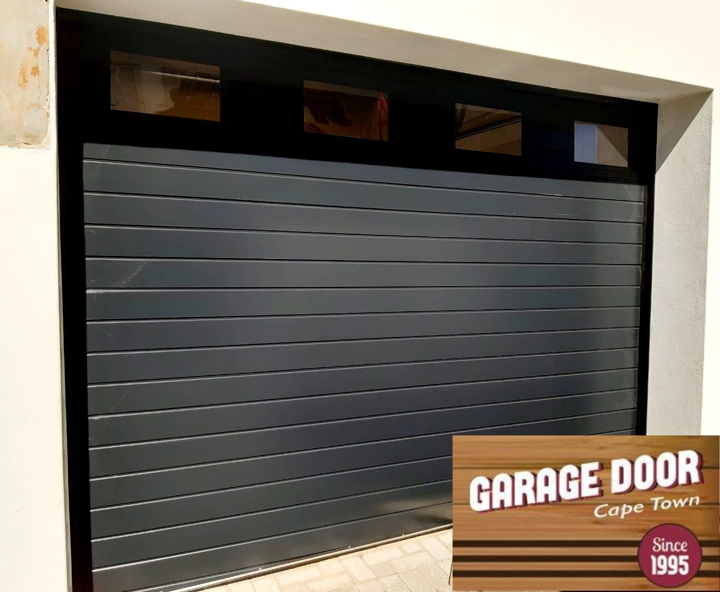 Dark gray horizontal slat garage door with small windows above. Cape Town logo.