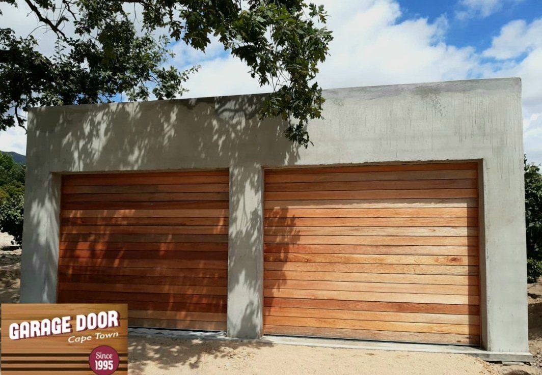 Two-car garage with horizontal wooden doors and a gray concrete exterior, under a blue sky with a tree.