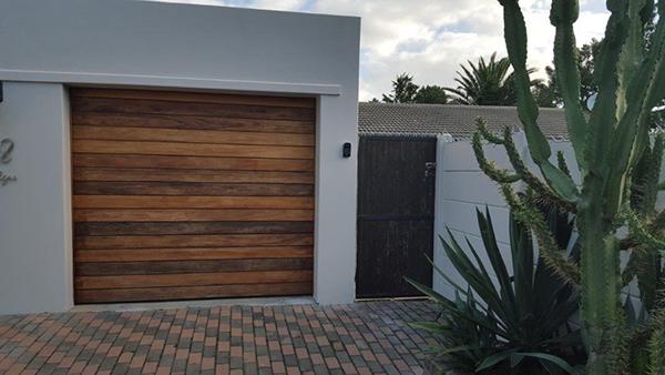 Wooden garage door and gate next to white wall and brick driveway, with cacti and greenery.