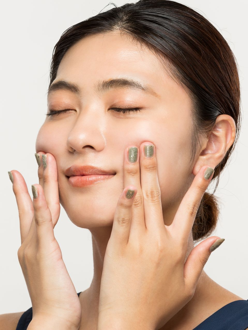 Woman with eyes closed, applying lotion to cheeks. White background, dark hair, blue top.