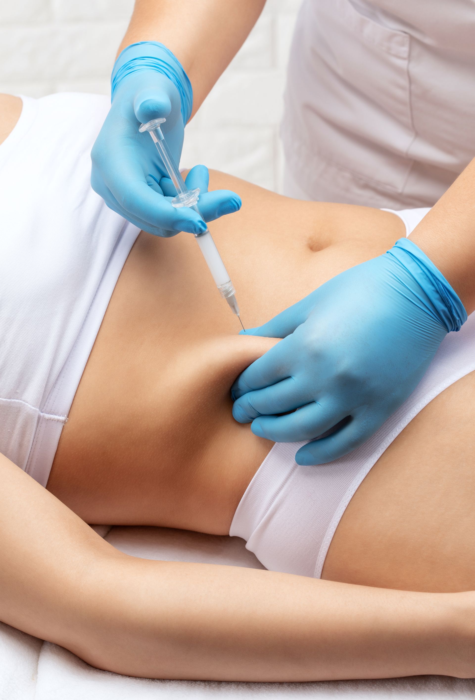 Hands in blue gloves injecting a syringe into a person's abdomen, on a white surface.