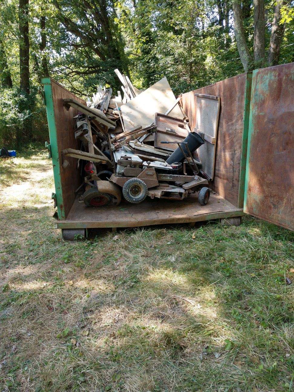Metal scrap in a large Yard Works Plus outdoors on grass, surrounded by trees.