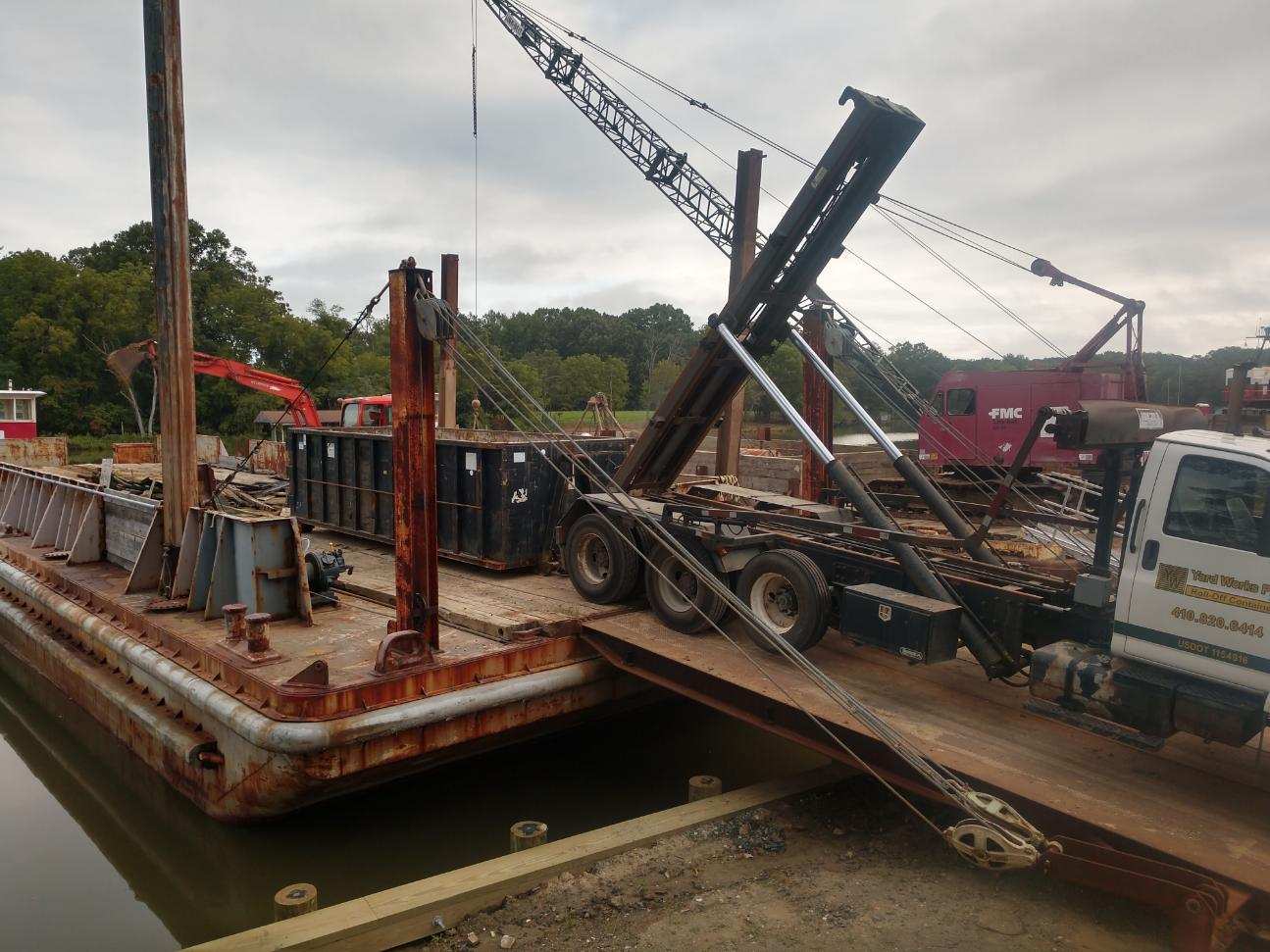 Yard Works Plus truck dumpster is on a barge, loading or unloading materials. marine construction site.
