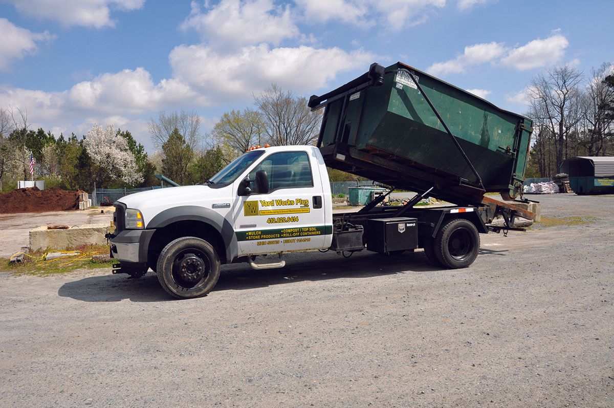 Yard Works Plus dumpster truck with a green dumpster on a gravel lot under a partly cloudy sky.