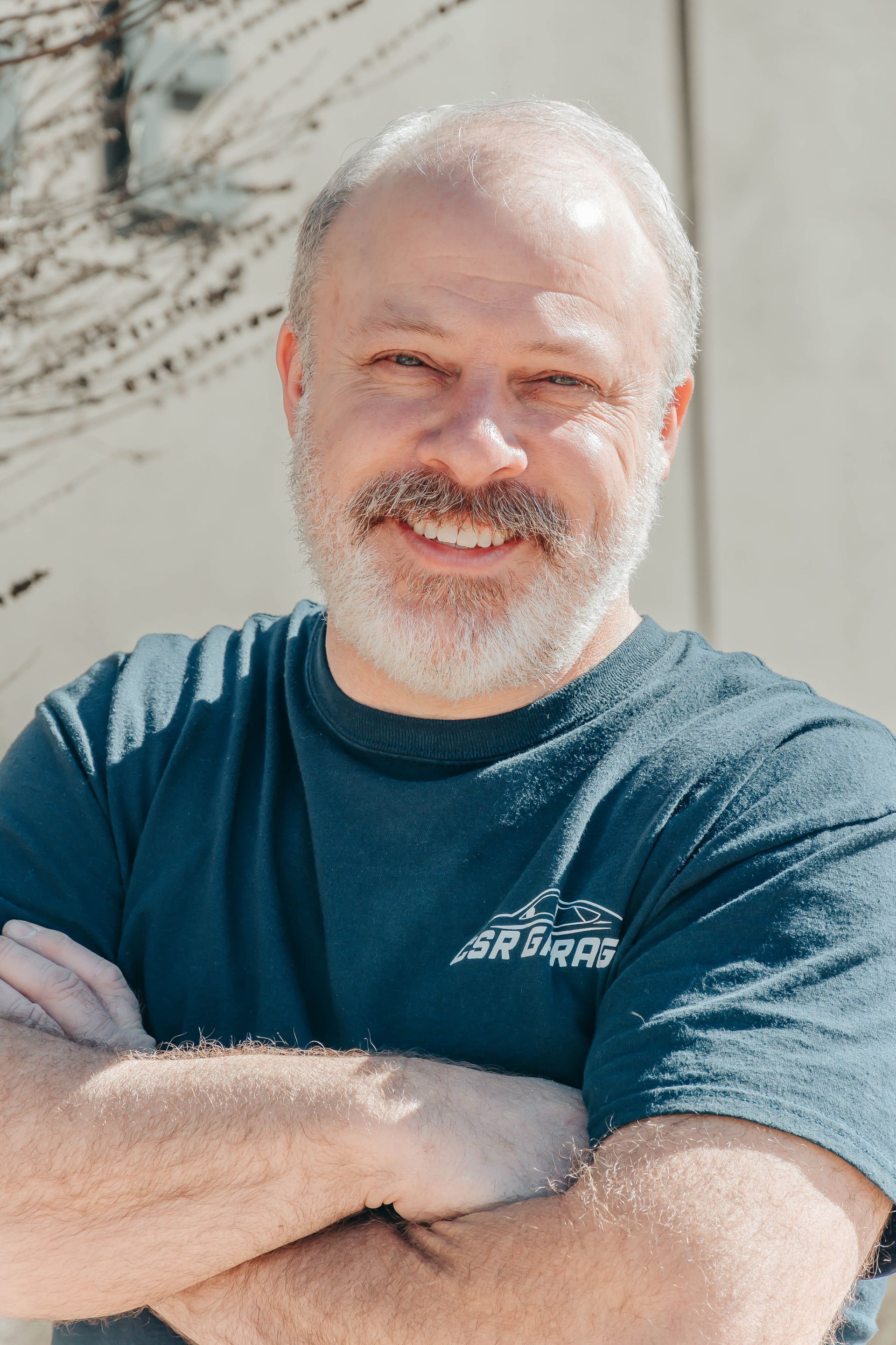 Smiling man in a teal T-shirt with arms crossed outdoors in bright sunlight