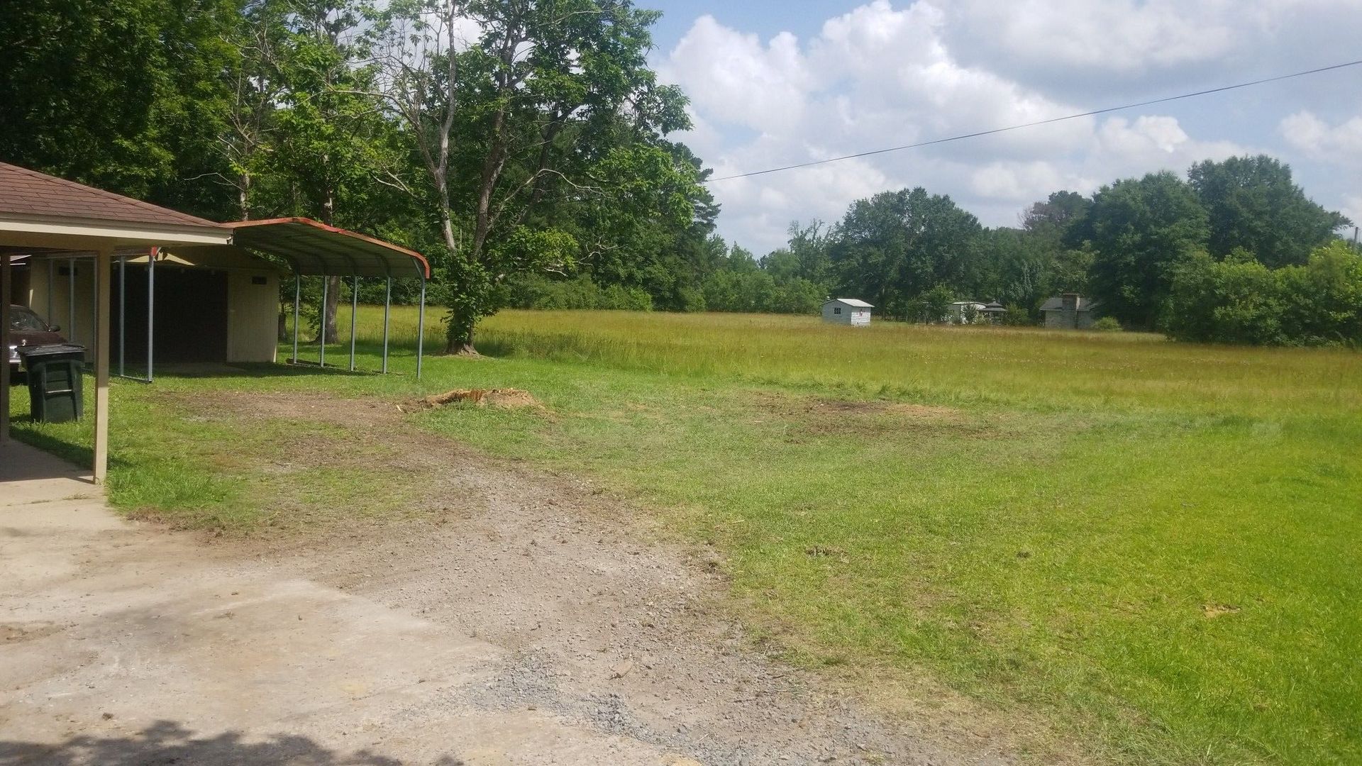 Grassy yard with a house on the left. A covered shelter and trees are visible. Sunny day.