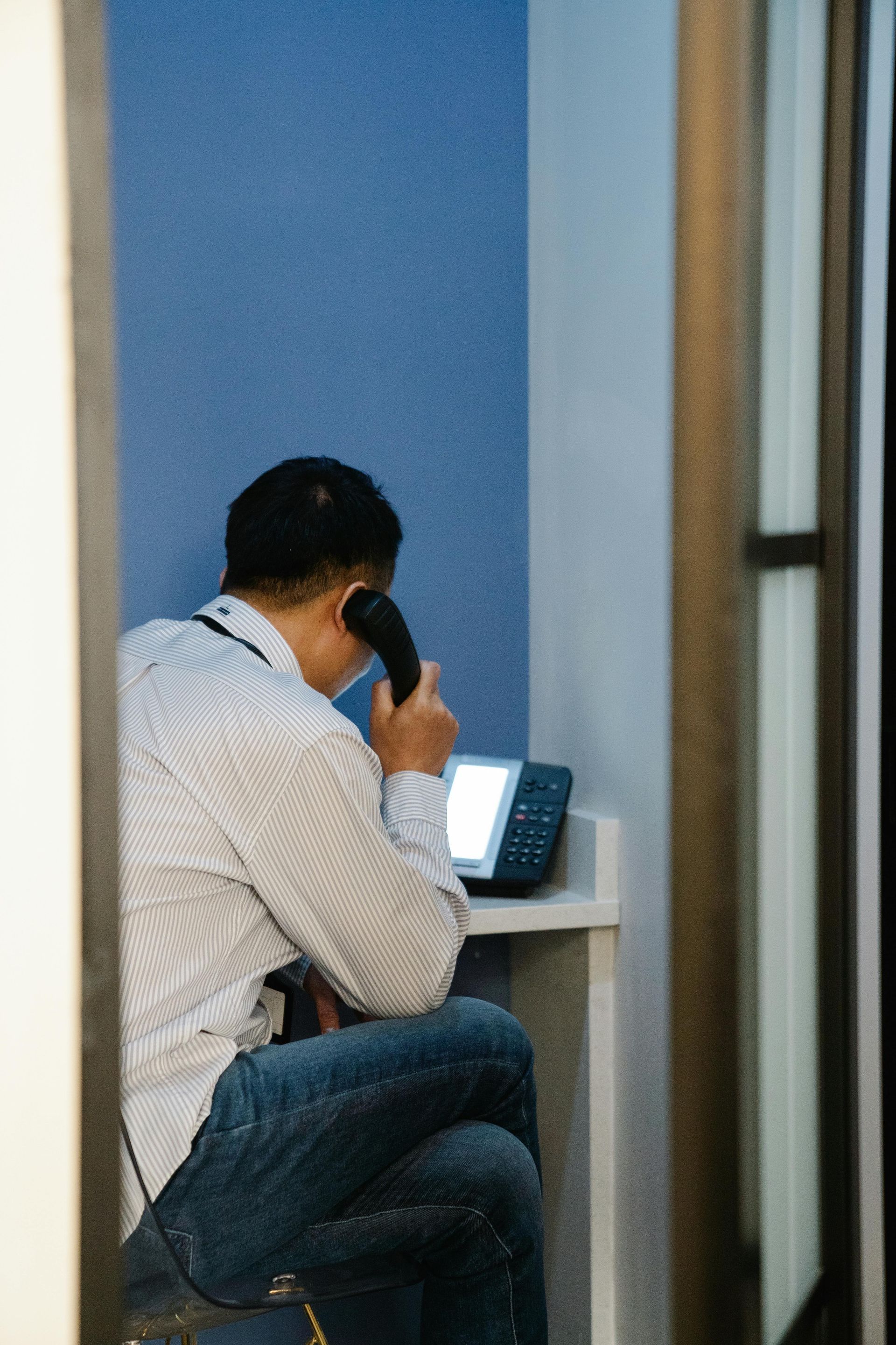 A person in a light shirt and blue jeans sits in a small booth, holding a telephone receiver to their ear.
