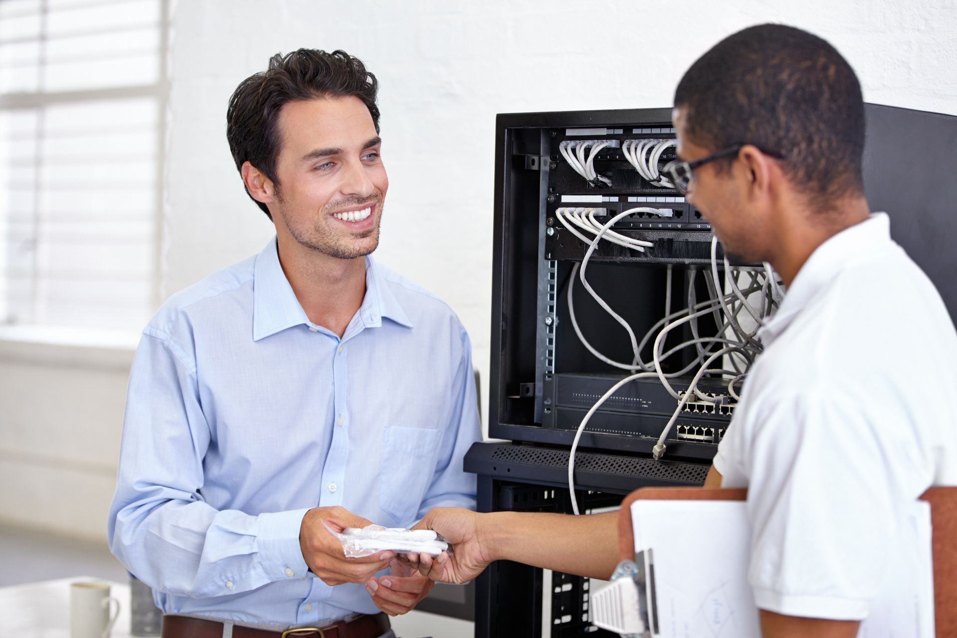 Two men by server rack, one offers white connectors, smiling.