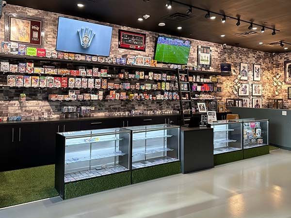 Inside a card shop, shelves stocked with sports cards; displays; televisions; brick accent wall.