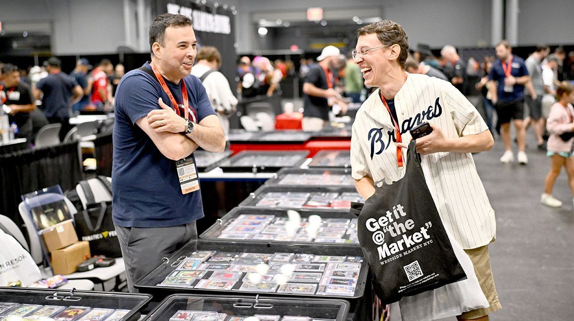 Two men are standing in front of a table at a convention talking to each other.