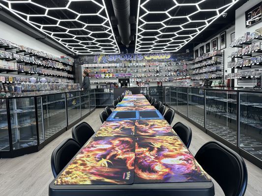 Interior of a card shop with playing tables, black chairs, and display cases filled with cards. Hexagonal ceiling lights.
