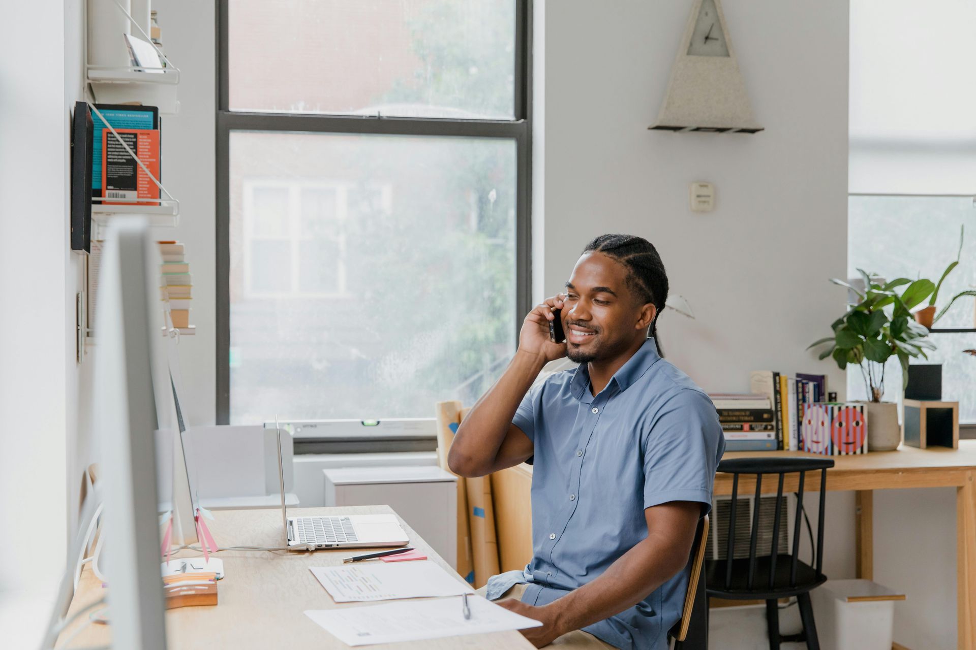 A person in a blue button-down shirt smiles while talking on a phone at a desk in a bright, modern office.