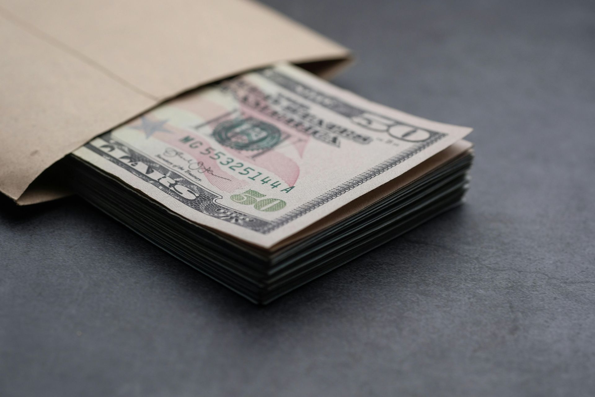 Stack of fifty-dollar bills in a brown envelope on a dark surface.