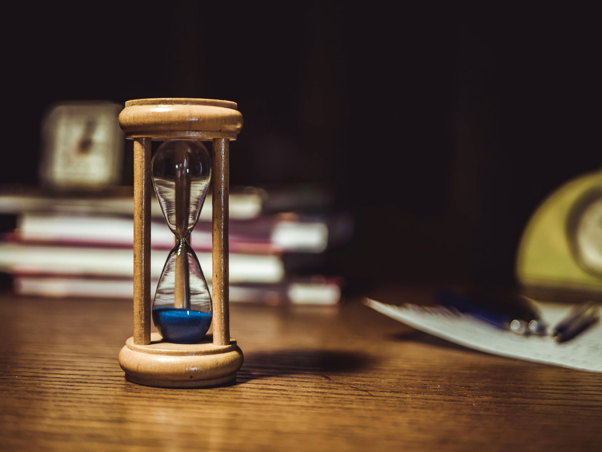 A wooden hourglass with blue sand sits on a wooden desk, with a stack of books and a blurred object in the background.