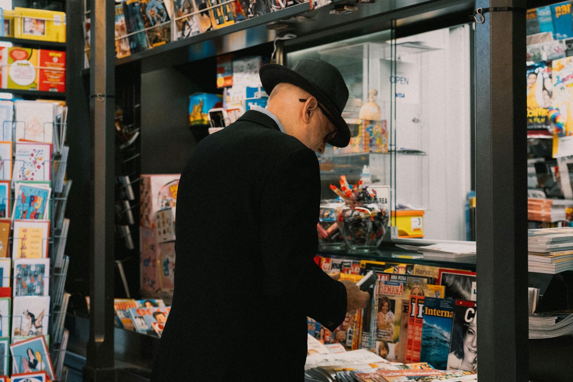 A person in a black coat and hat browsing magazines at a small shop counter surrounded by shelves of merchandise.