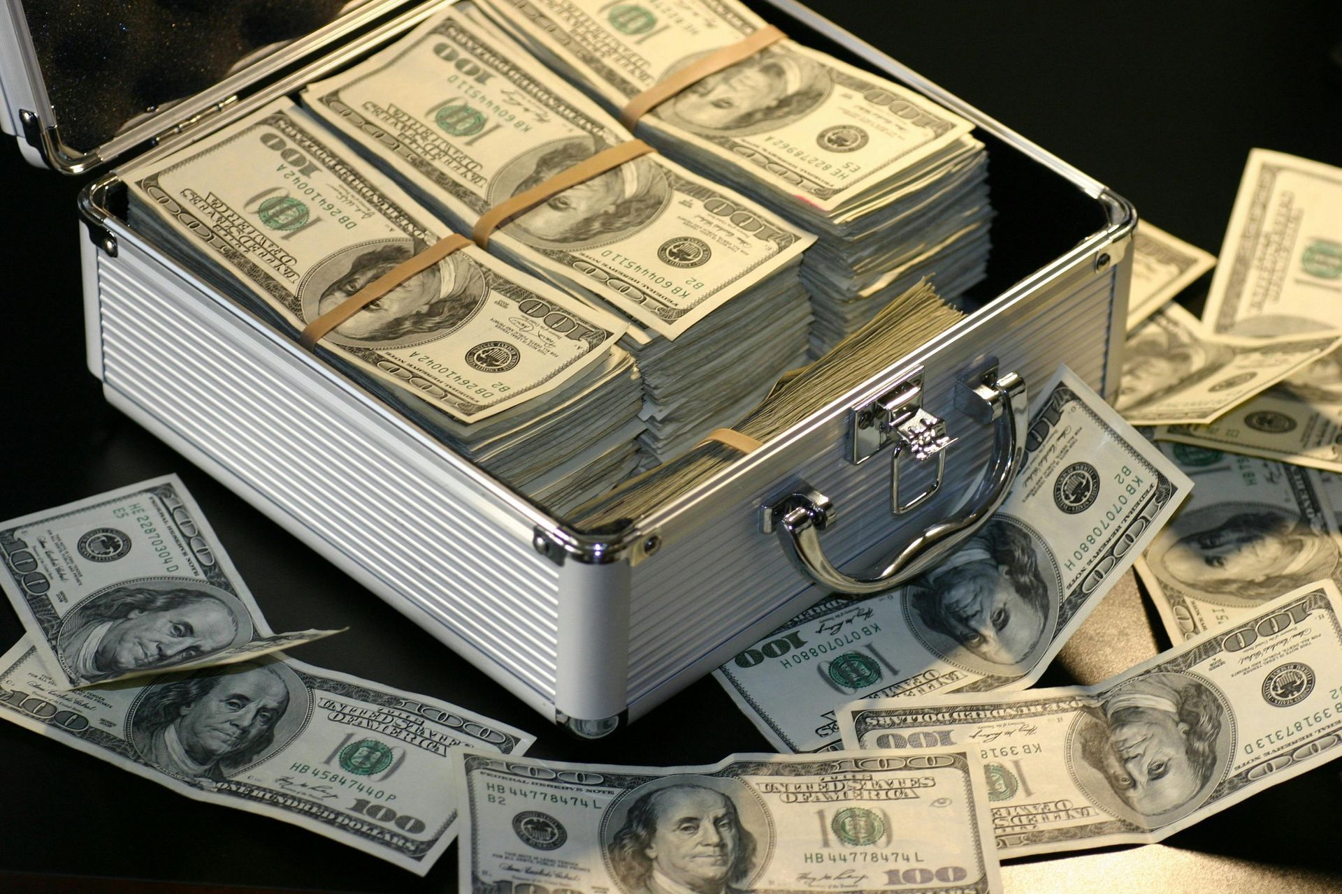 Suitcase overflowing with stacks of $100 bills; currency scattered around on dark surface.