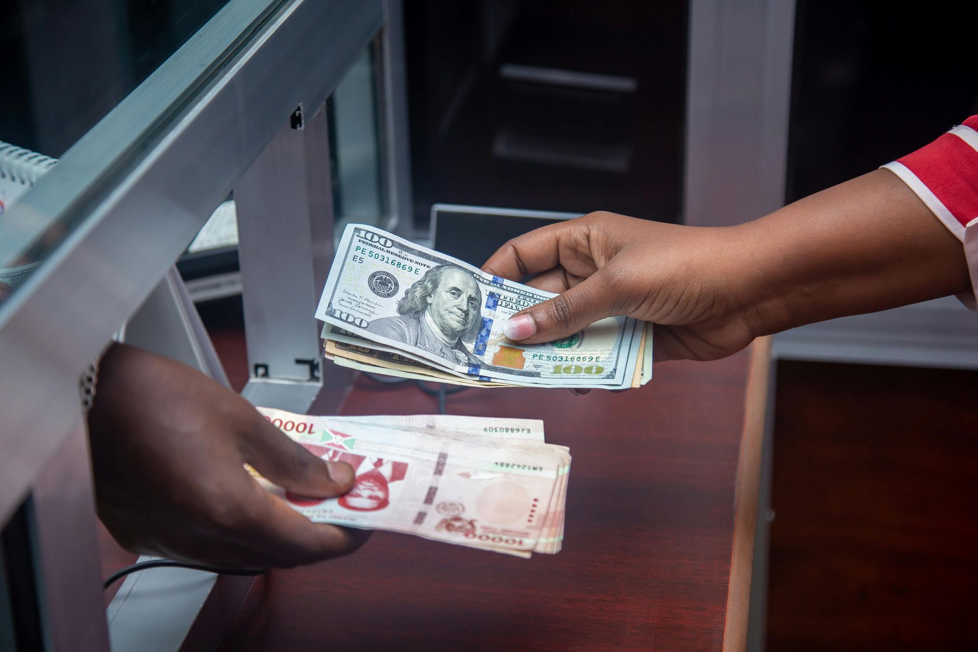 Hands exchanging US dollars and other paper currency across a bank teller counter.
