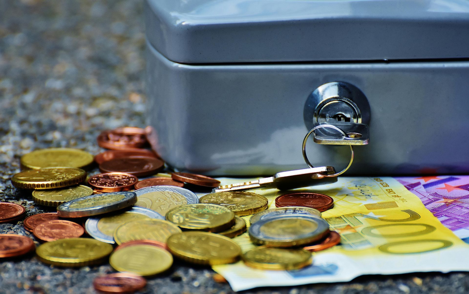 A grey metal cash box with a key inserted, resting on a pile of scattered Euro coins and a 200 Euro banknote.