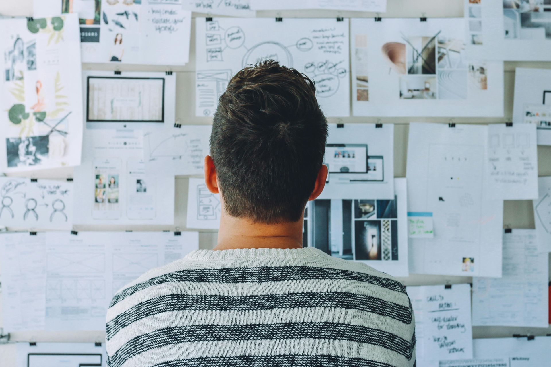 Person in striped sweater looking at a wall covered with sketches, diagrams, and notes.