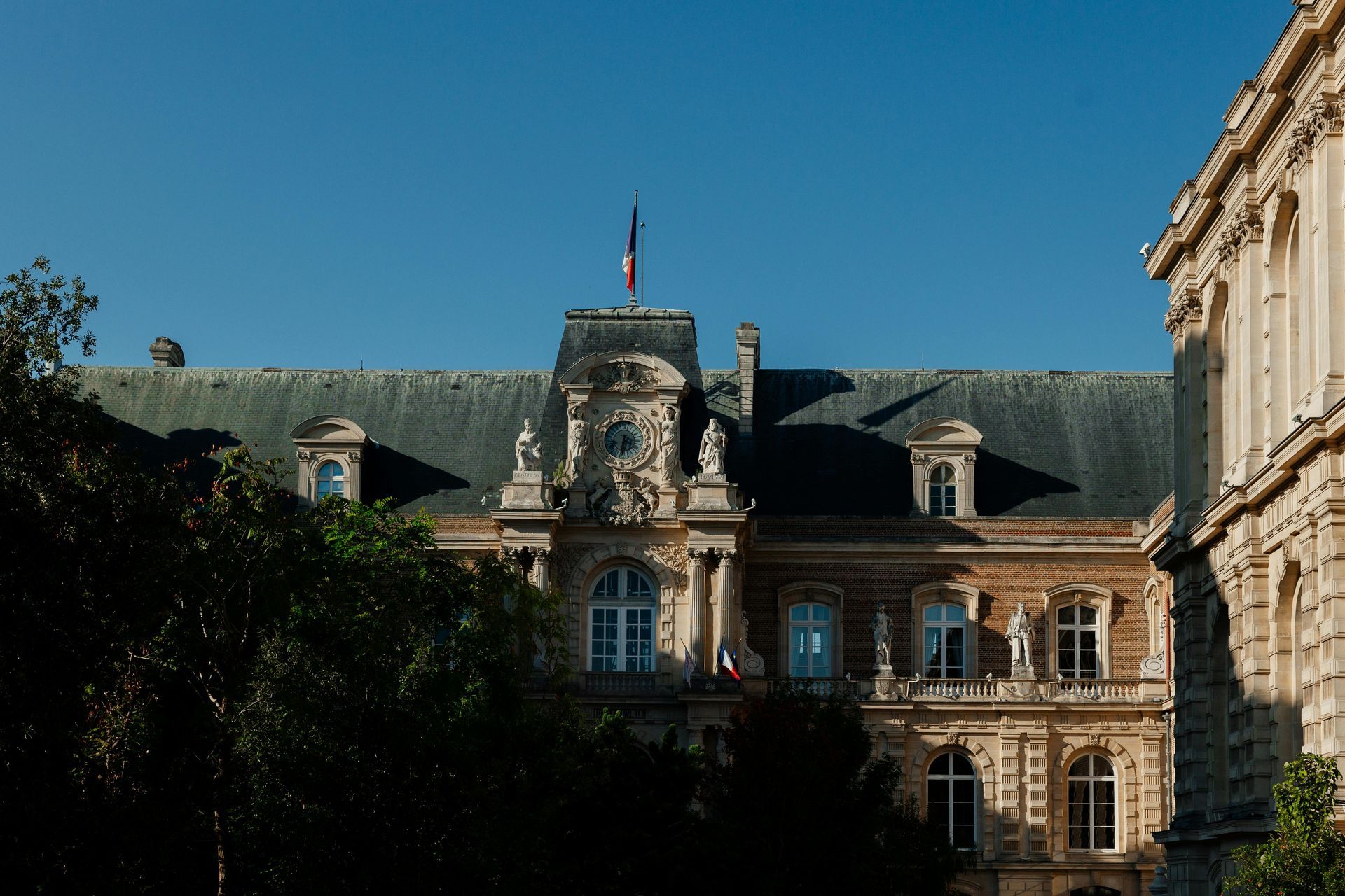 Classical French architecture with a central dormer, stone facades, ornate sculptures, and a flag against a blue sky.
