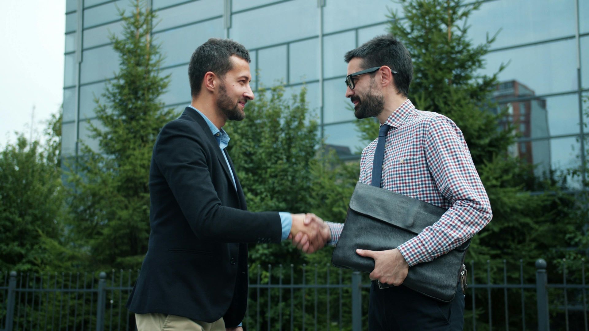 Two business professionals shaking hands in front of a modern glass office building.