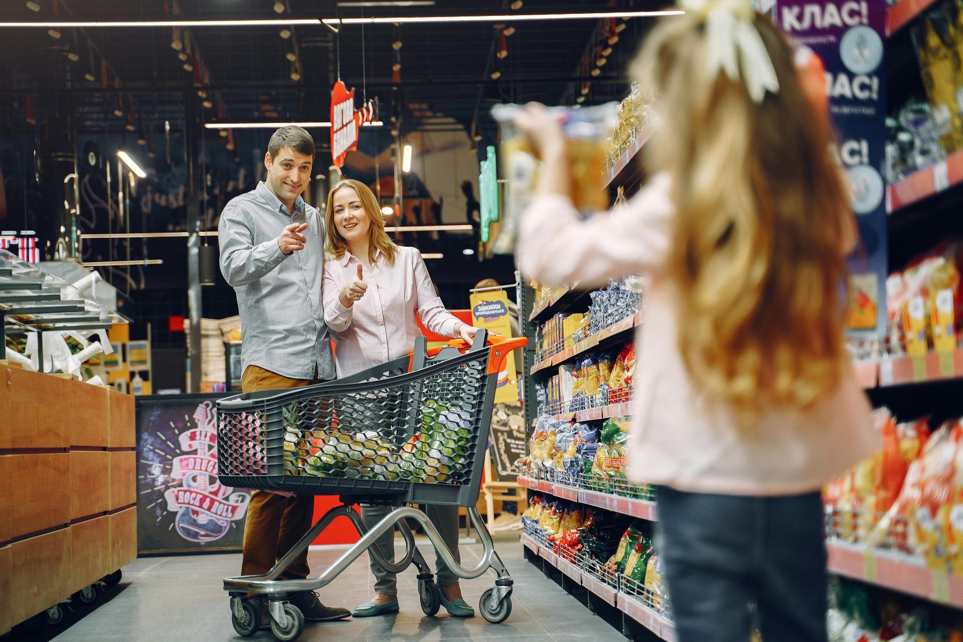 Family shopping at grocery store; daughter holding snacks, parents smiling, pushing cart.