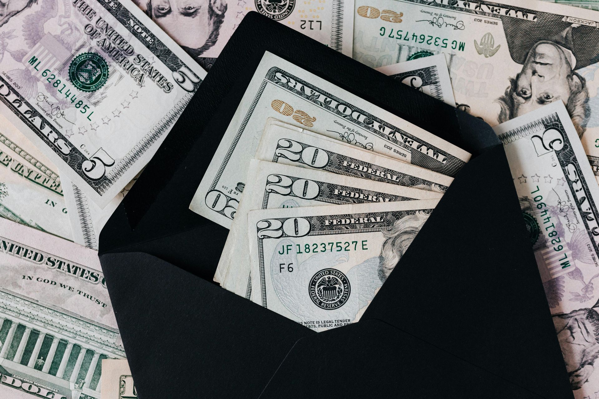 A black envelope filled with twenty-dollar bills, surrounded by scattered five-dollar US bills on a light surface.