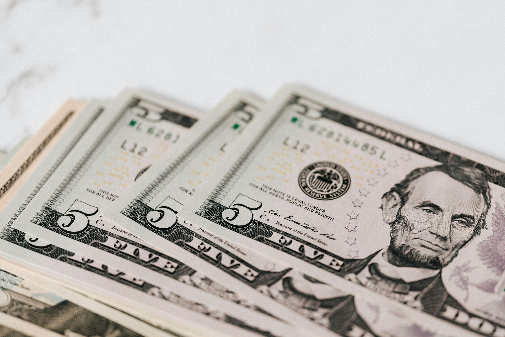 A close-up, angled view of a stack of several US five-dollar bills showing Abraham Lincoln's portrait.