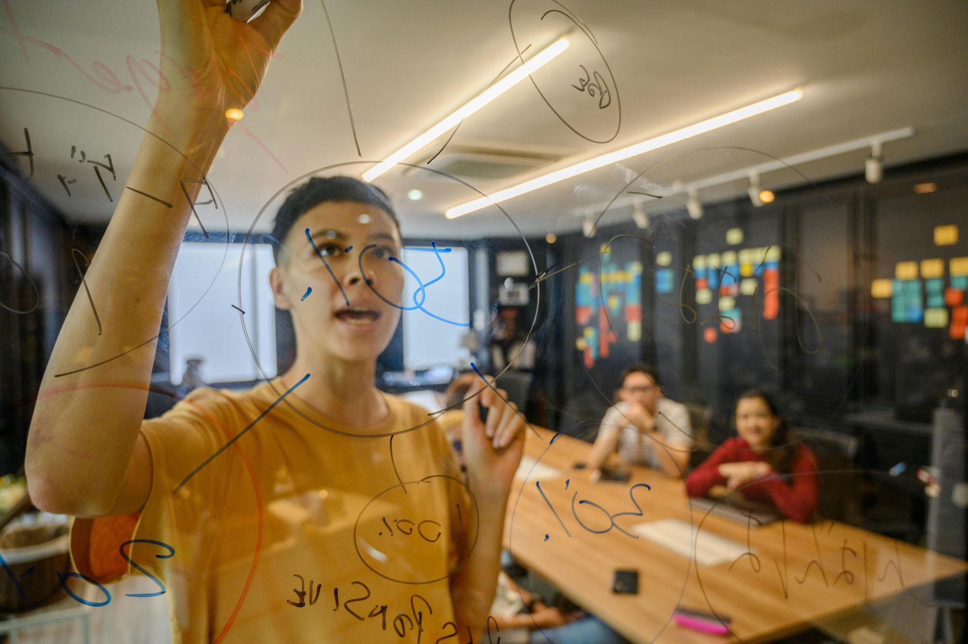 A person writes on a glass office wall during a meeting, while two others watch from a conference table.