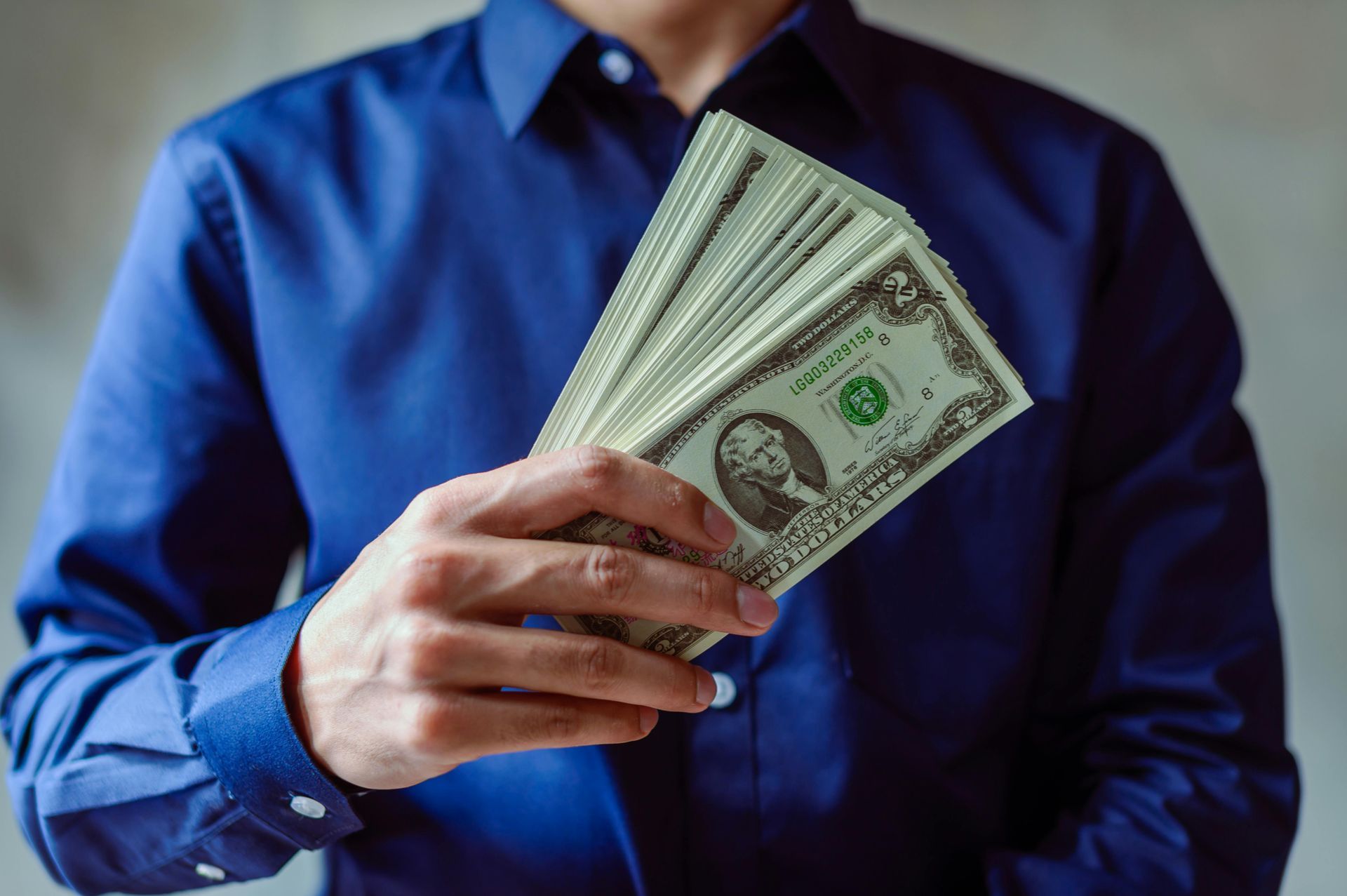 A person in a blue button-down shirt holds a fan of US one-dollar bills.