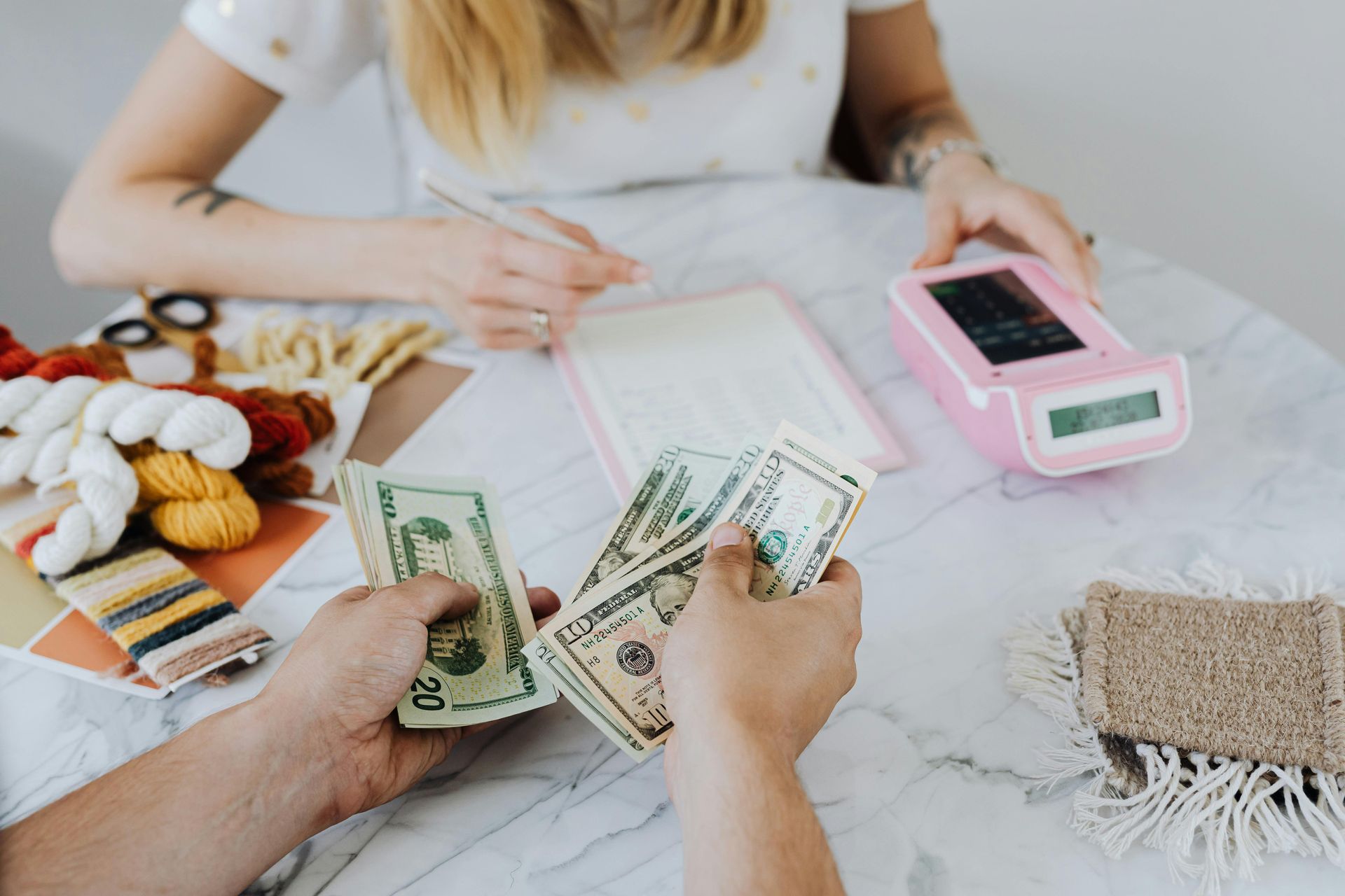Two sets of hands exchange US cash over a table containing yarn supplies, a notepad, and a pink digital scale.