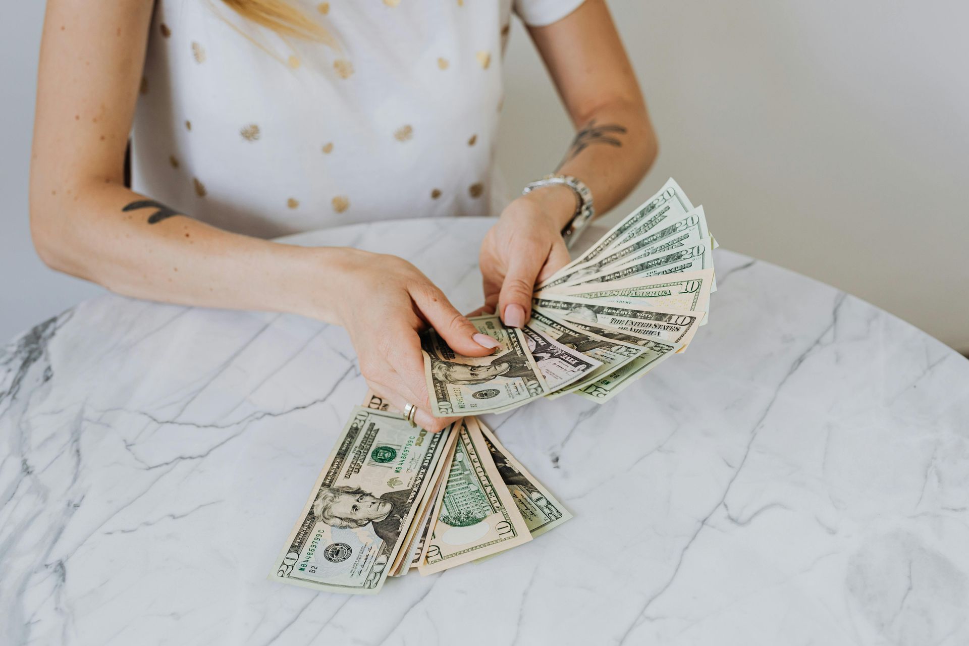 A person holds and fans out multiple US twenty-dollar bills over a white marble table.