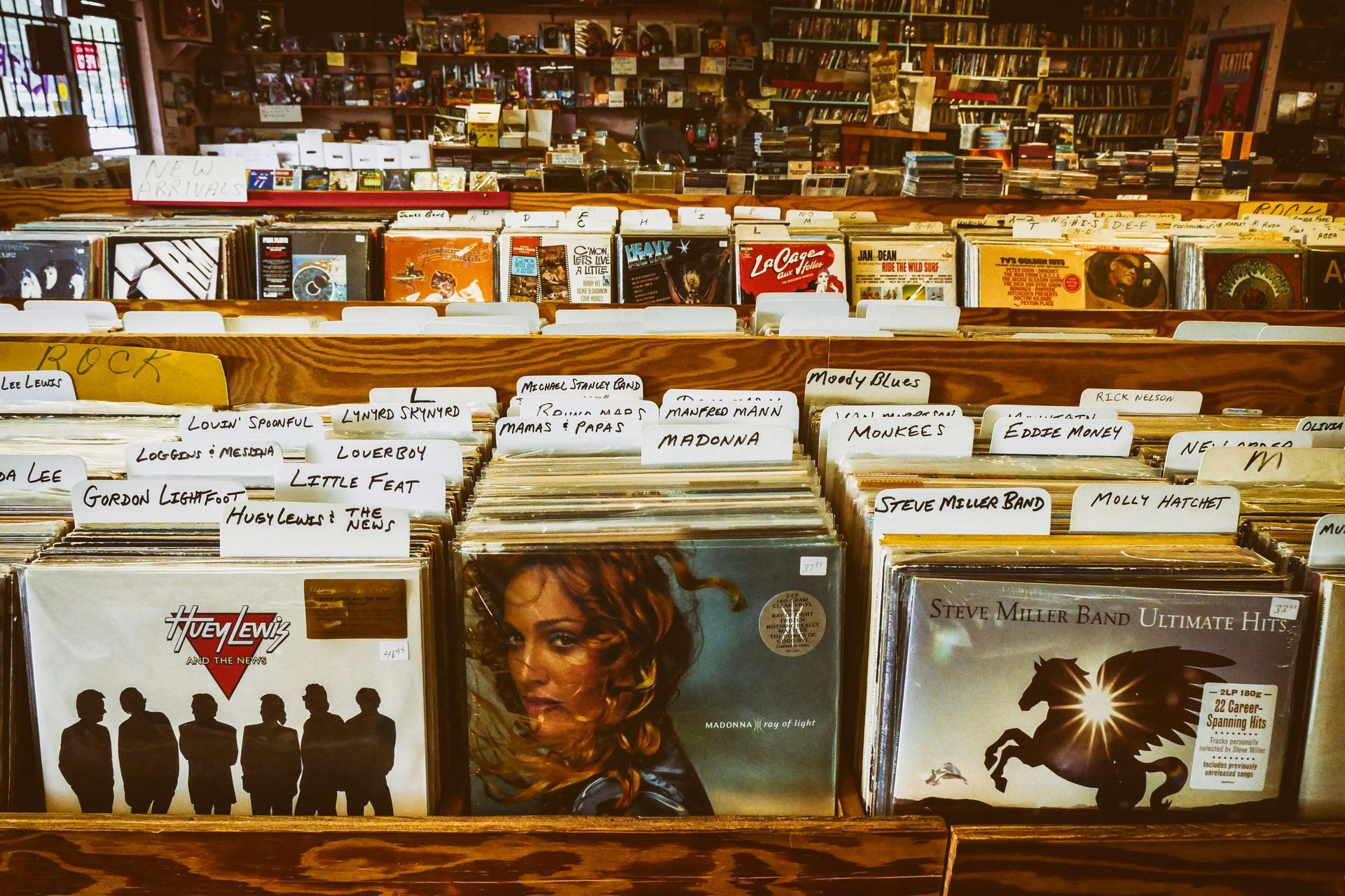 Record store, close-up of vinyl albums in wooden bins. Various album covers, titles on dividers, store shelves in background.