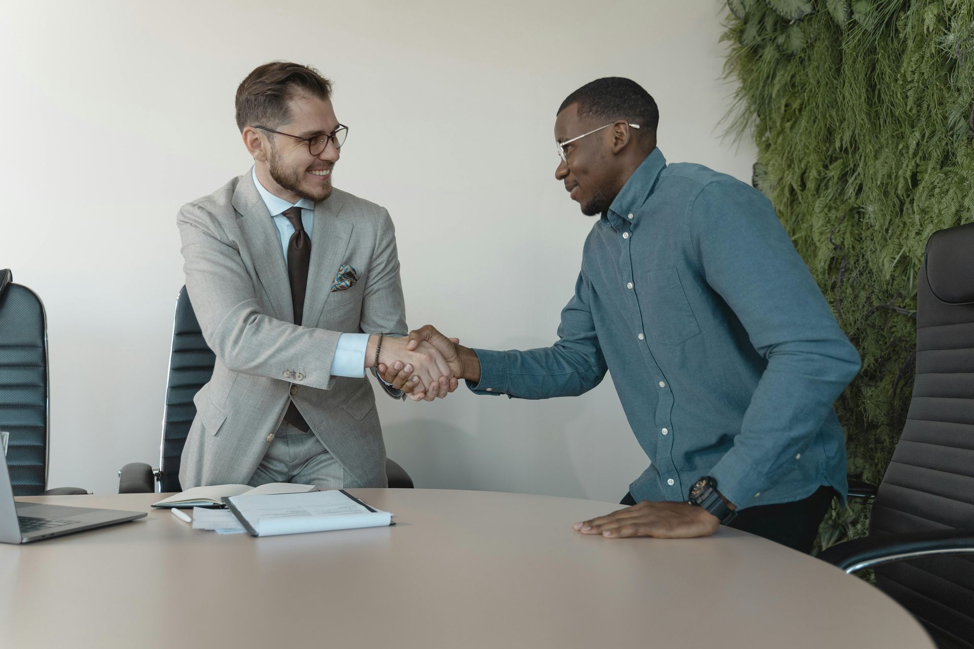 Two people in a professional office setting shake hands across a conference table.